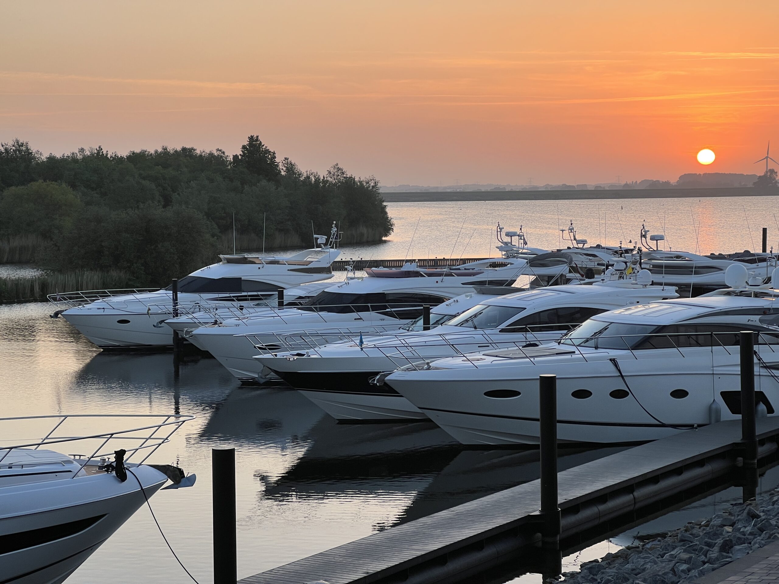 Zonsopgang boven een jachthaven met luxe jachten aan een steiger en bomen op de achtergrond.
