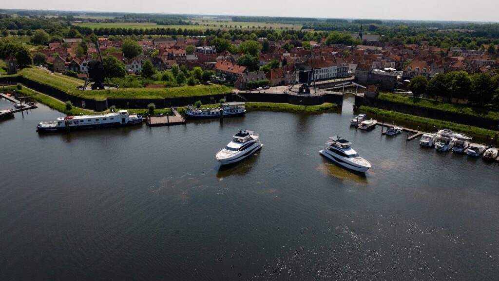Luchtfoto van twee luxe jachten van Princess Yachts in een historische haven met op de achtergrond een pittoresk stadje en groene landschappen.