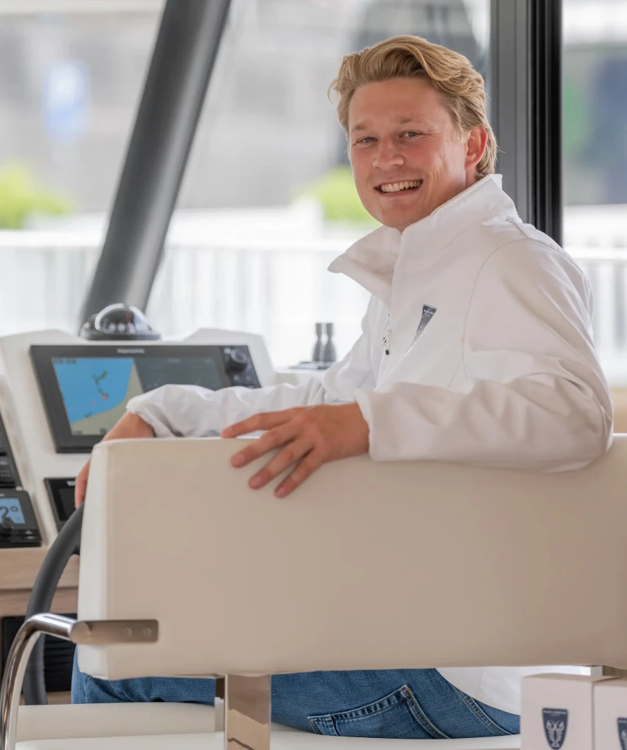 Een man in een witte jas glimlacht terwijl hij achter het stuur van een jacht zit, met een navigatiescherm op de achtergrond.