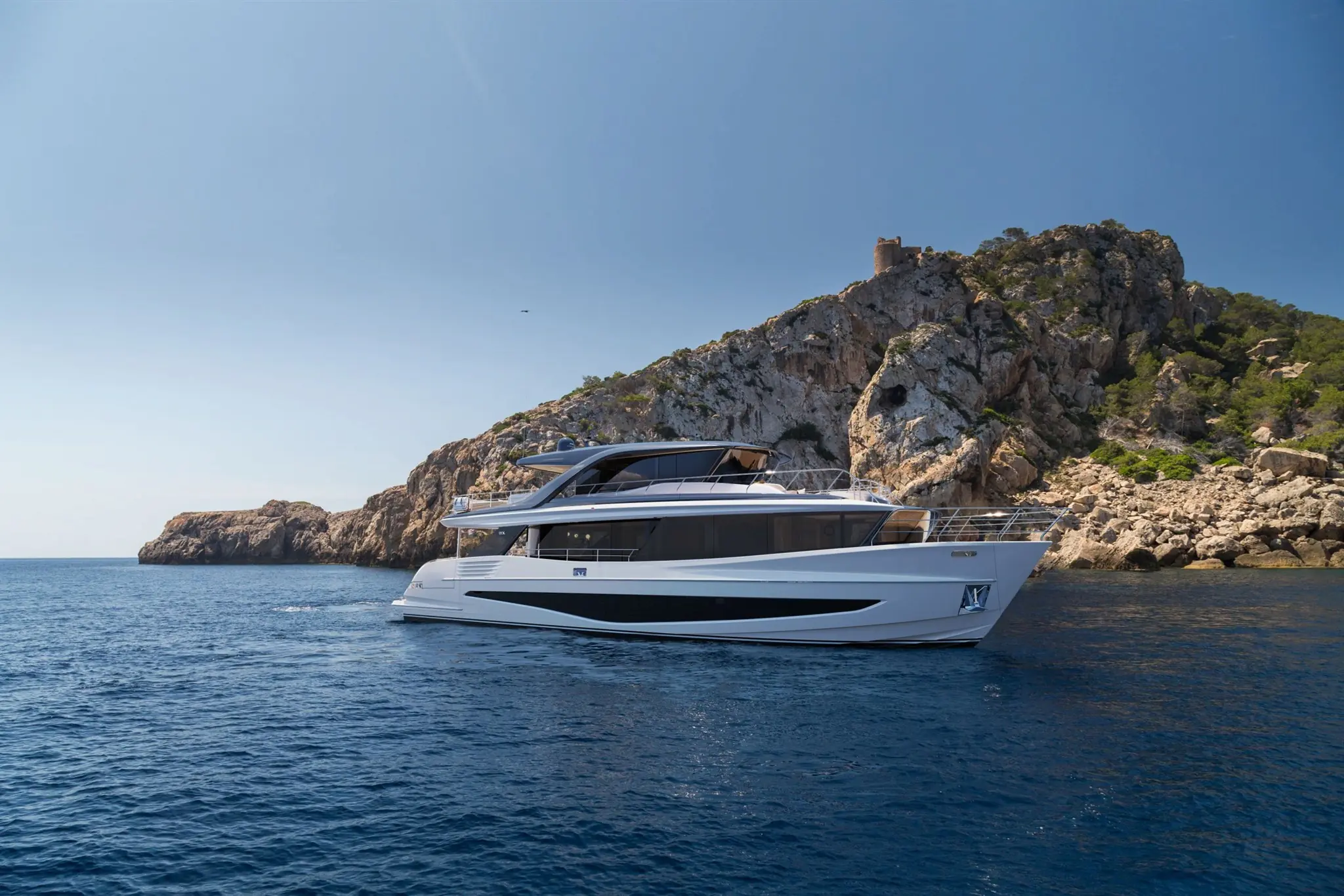 Een luxe Princess Yachts X Class vaart langs een rotsachtig kustlandschap onder een heldere blauwe hemel.